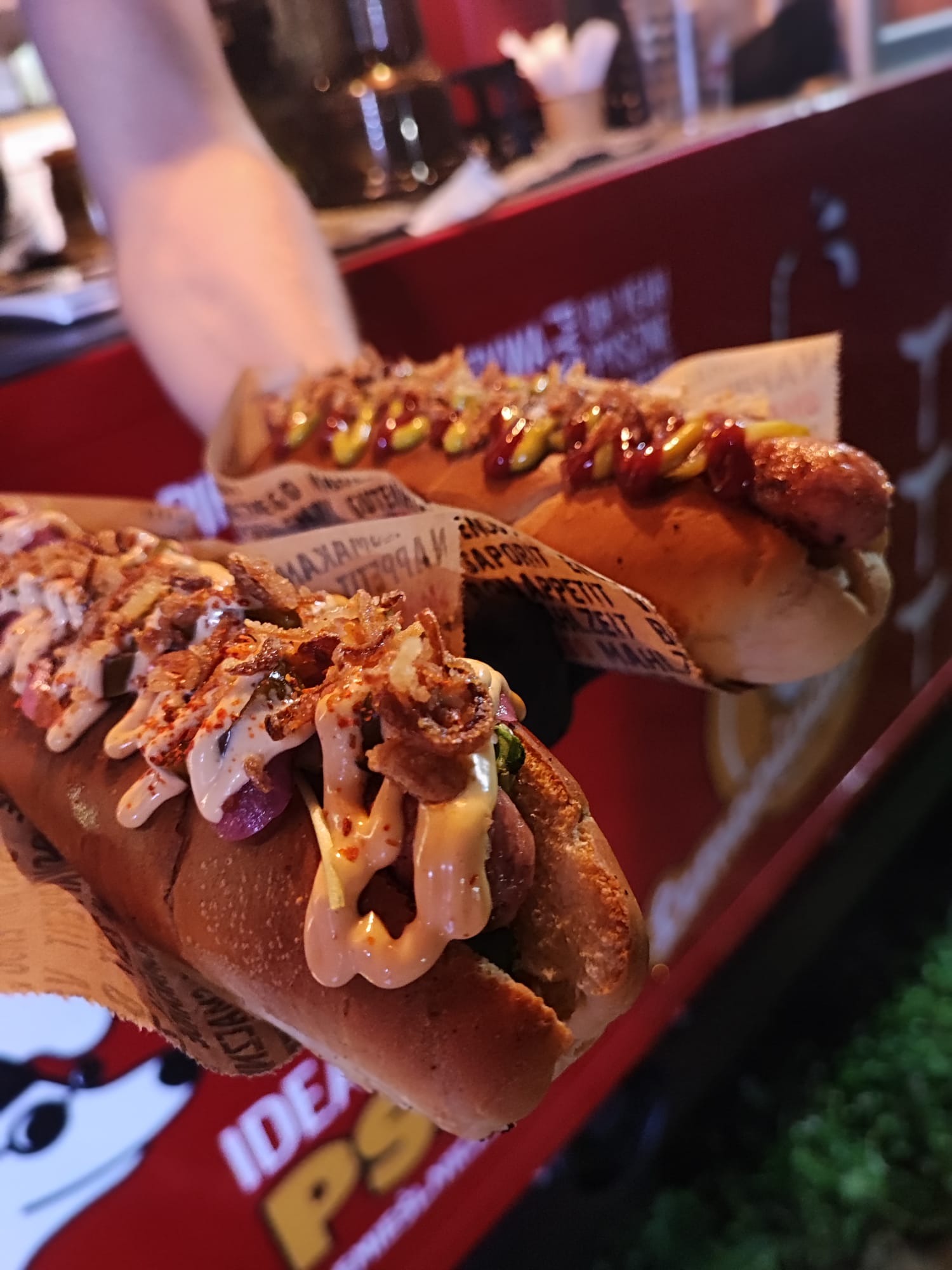 Dwa hot dogi z różnymi sosami i posypkami, podane na papierowych podkładkach z nadrukiem, trzymane w ręce na tle czerwonej tacy i ciemnego otoczenia.