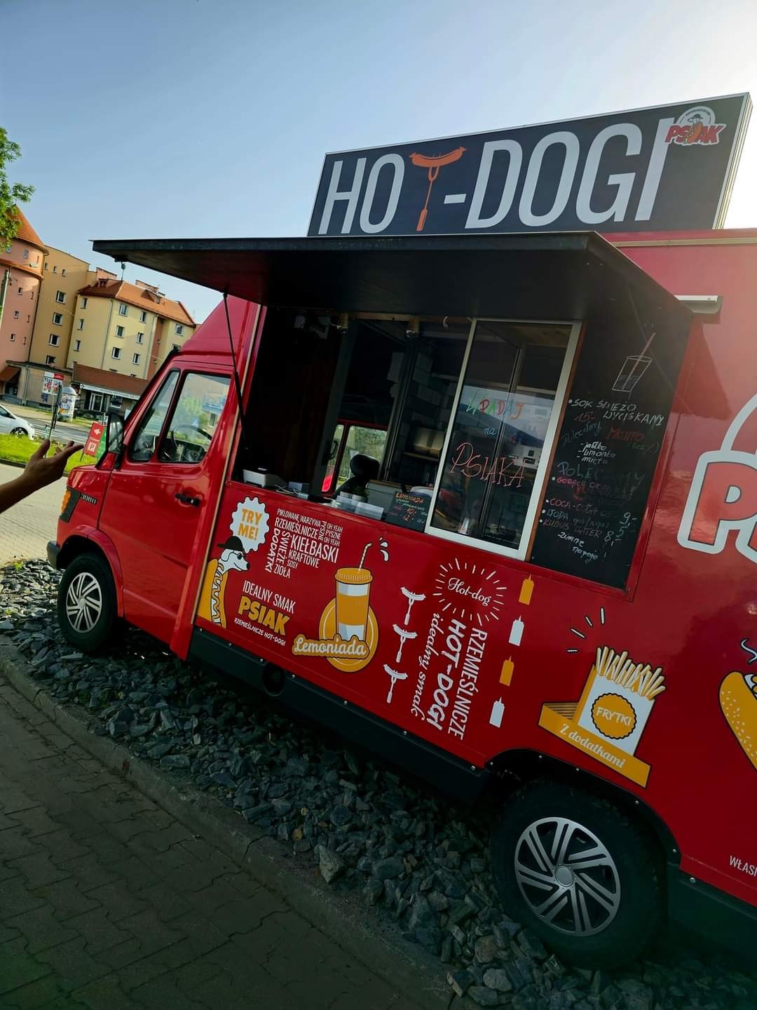 Czerwony food truck z szyldem 'HOT-DOGI', zaparkowany na żwirowym podłożu, z menu wypisanym kredą na szybie i grafikami reklamującymi hot-dogi, lemoniadę i frytki.