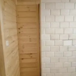 Ściana wykończona jasnym drewnem boazeryjnym sąsiadująca z fragmentem ściany wyłożonej białymi, błyszczącymi, prostokątnymi płytkami ceramicznymi, prawdopodobnie w trakcie prac wykończeniowych.