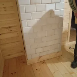 Wnętrze wykończone drewnem z fragmentem białej, kafelkowej konstrukcji przypominającej piec lub kominek, w trakcie prac wykończeniowych, widoczny but pracownika i młotek na podłodze.