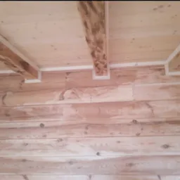 Drewniana boazeria ścienna i sufitowa z widocznymi belkami stropowymi w jasnym odcieniu, wykończona białymi listwami przypodłogowymi.