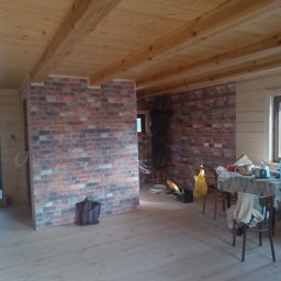 PerfectKam - Wnętrze drewnianego domu w trakcie remontu: ściana z cegły dekoracyjnej, drewniana podłoga i sufit, widoczny pracownik na stołku montujący element na ścianie.