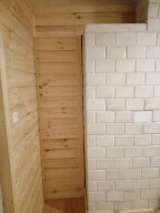 Ściana wykończona jasnym drewnem boazeryjnym sąsiadująca z fragmentem ściany wyłożonej białymi, błyszczącymi, prostokątnymi płytkami ceramicznymi, prawdopodobnie w trakcie prac wykończeniowych.