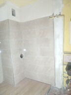 Wykończona kabina prysznicowa z beżowymi płytkami, widoczna bateria i fragment instalacji wodnej, sufit w odcieniu żółci, ściana częściowo przygotowana do położenia płytek.