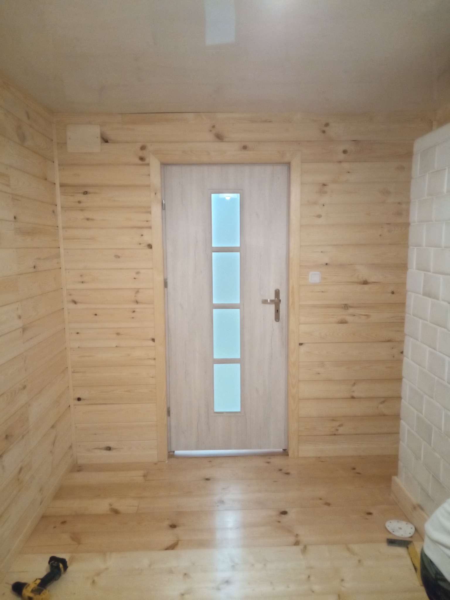 Wnętrze wykończone boazerią z jasnego drewna, z widoczną częściowo otwartą białą ścianą z cegiełek po prawej stronie i białą, częściowo przeszkloną stolarką drzwiową na wprost, w dolnym lewym rogu...