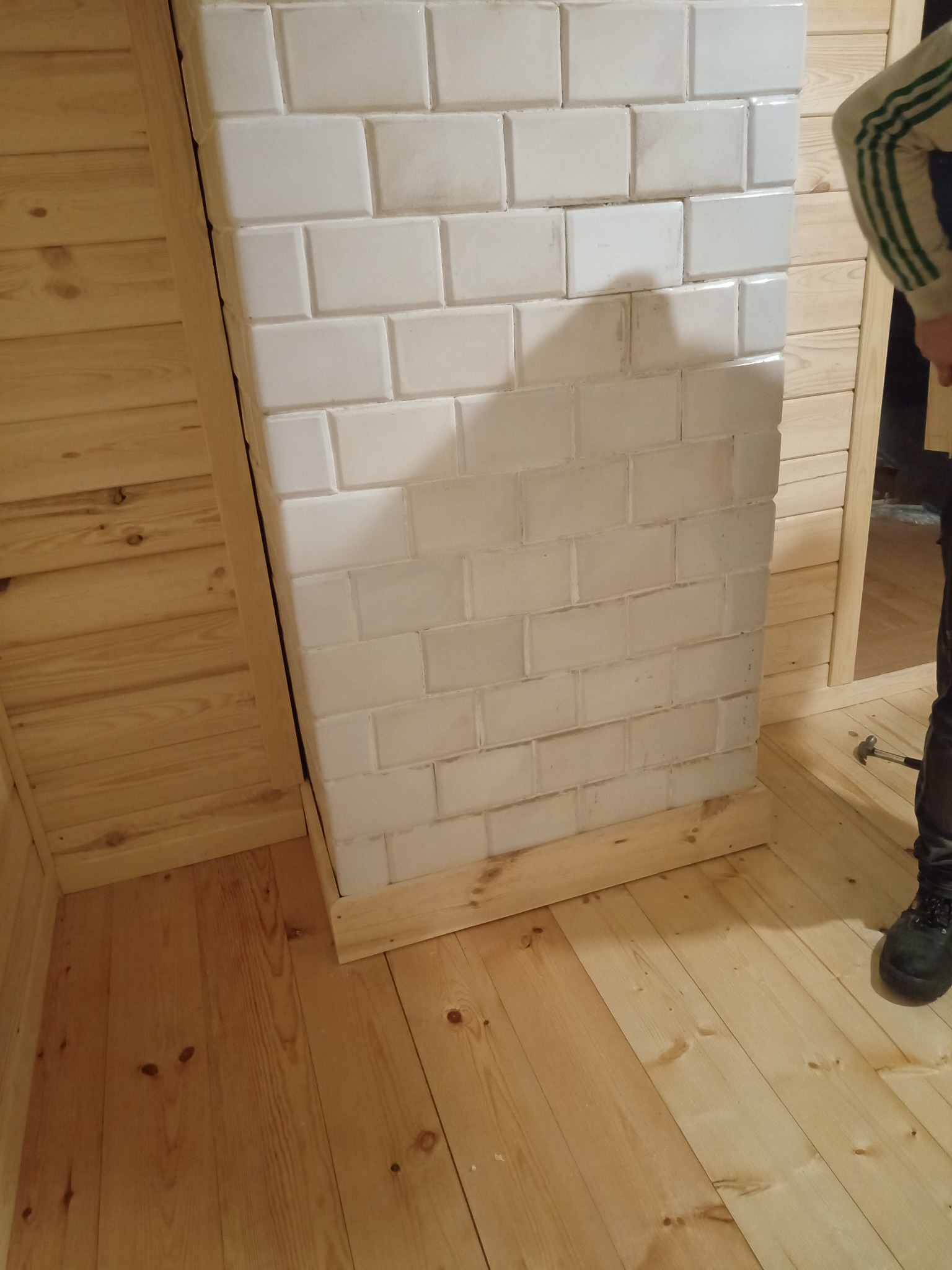 Wnętrze wykończone drewnem z fragmentem białej, kafelkowej konstrukcji przypominającej piec lub kominek, w trakcie prac wykończeniowych, widoczny but pracownika i młotek na podłodze.