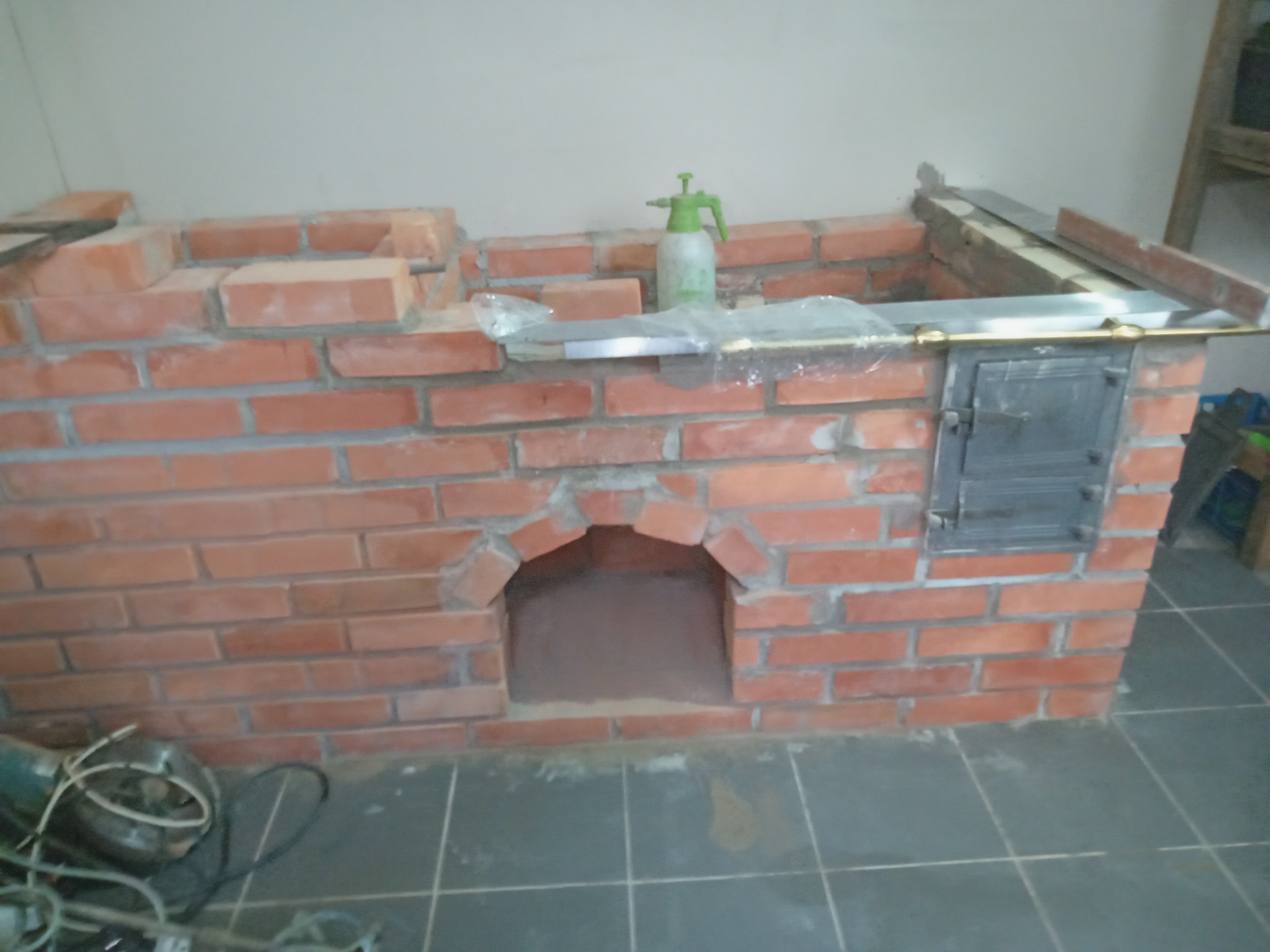 Wnętrze w trakcie budowy pieca z czerwonej cegły z otworem paleniskowym i metalowymi drzwiczkami, na wierzchu poziomnica, spryskiwacz i folia ochronna, widoczne narzędzia na podłodze.