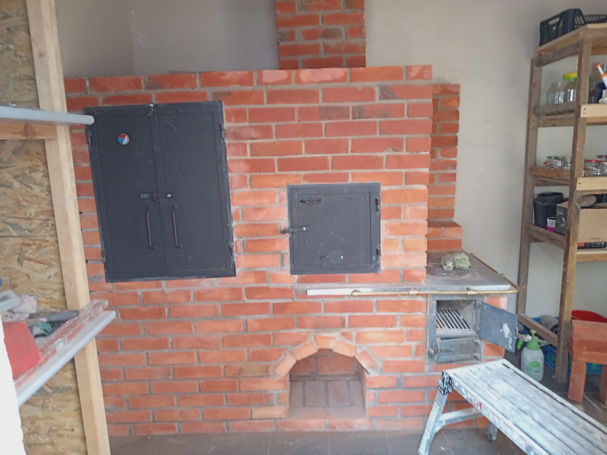 Murowany piec kuchenny z cegły z trzema czarnymi, żeliwnymi drzwiczkami, widoczny fragment komina, drabina malarska i drewniany regał z słoikami w tle.