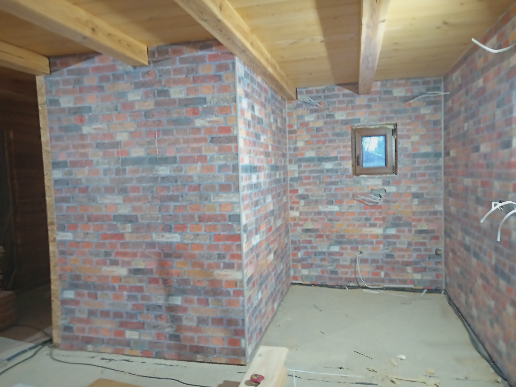 Fragment pokoju z wykończonymi ścianami z cegły dekoracyjnej, drewnianym stropem i wystającym okienkiem. Widoczne kable elektryczne wystające ze ścian.