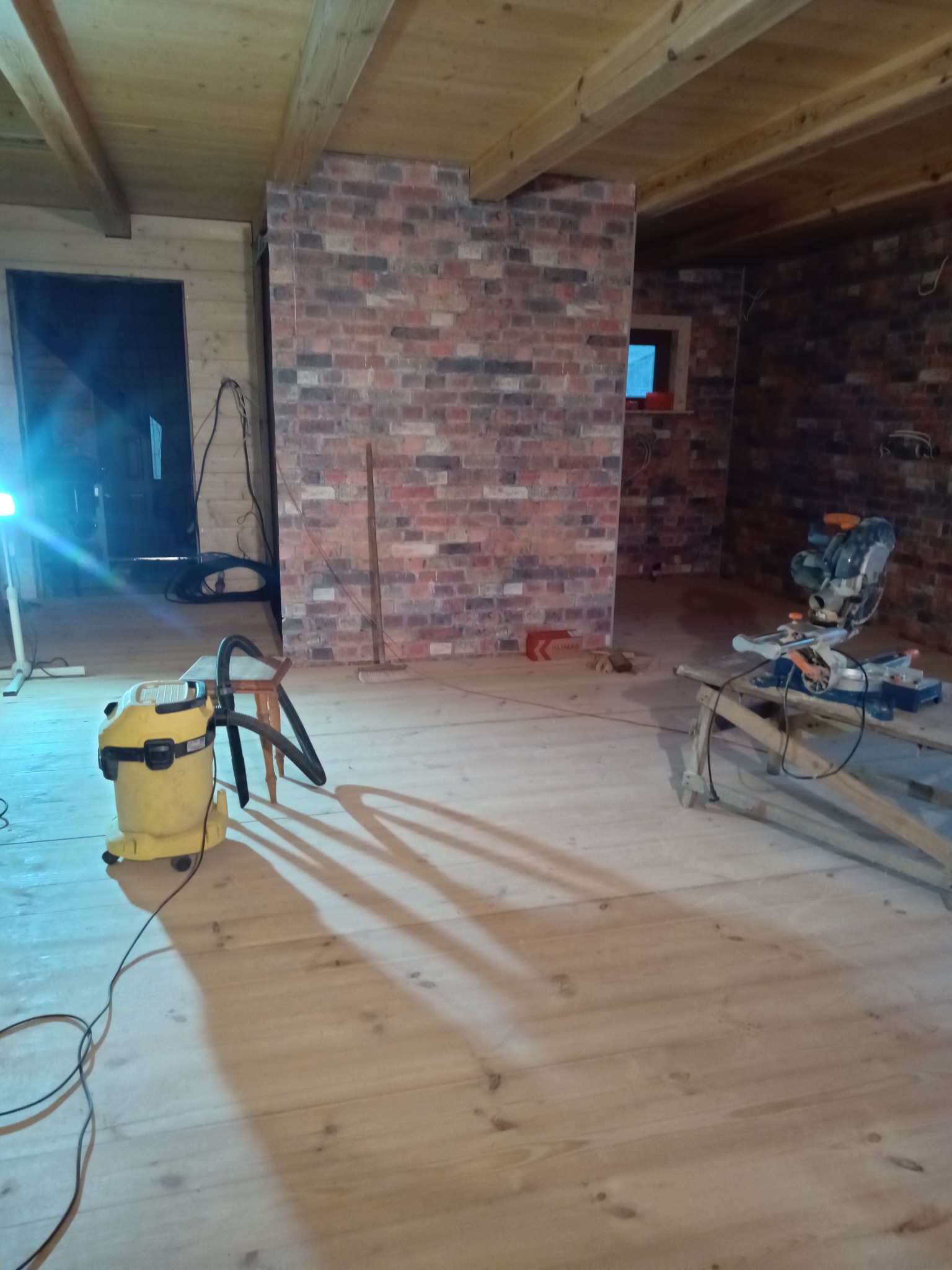 Wnętrze w trakcie remontu: drewniany sufit i podłoga, fragment ściany z cegły, odkurzacz przemysłowy i piła ukośnica na drewnianym stojaku, widoczne przewody i narzędzia.
