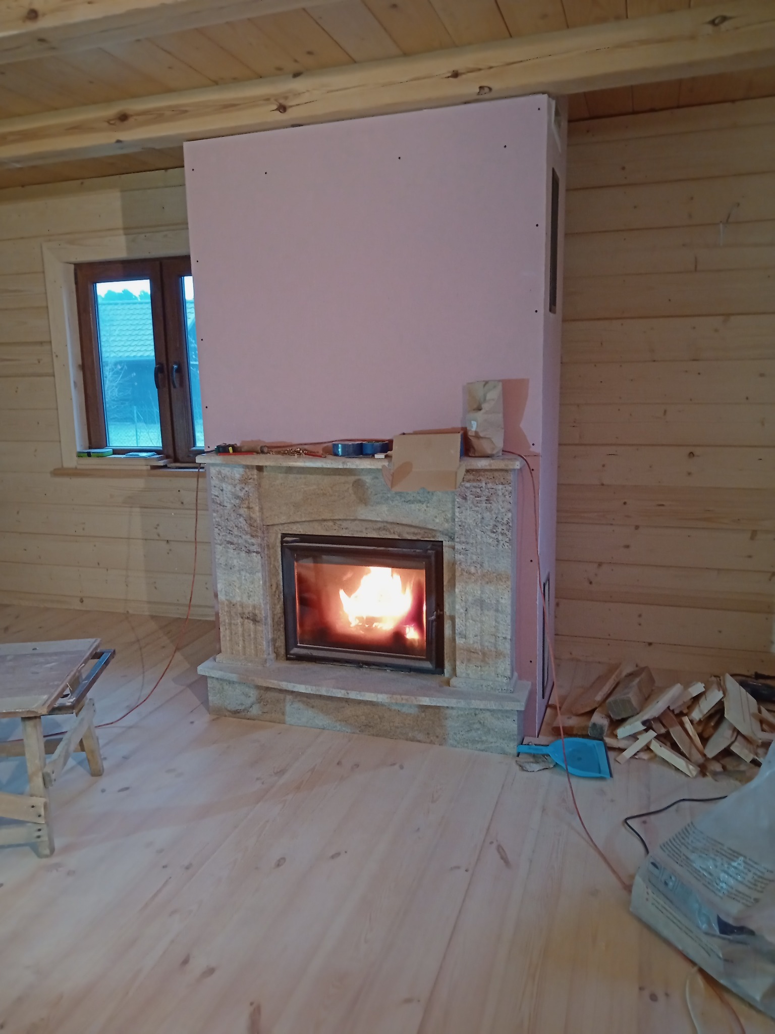 Kominek z obudową z kamienia i płyt karton-gips, w trakcie montażu w drewnianym domu, widoczne narzędzia i materiały budowlane.