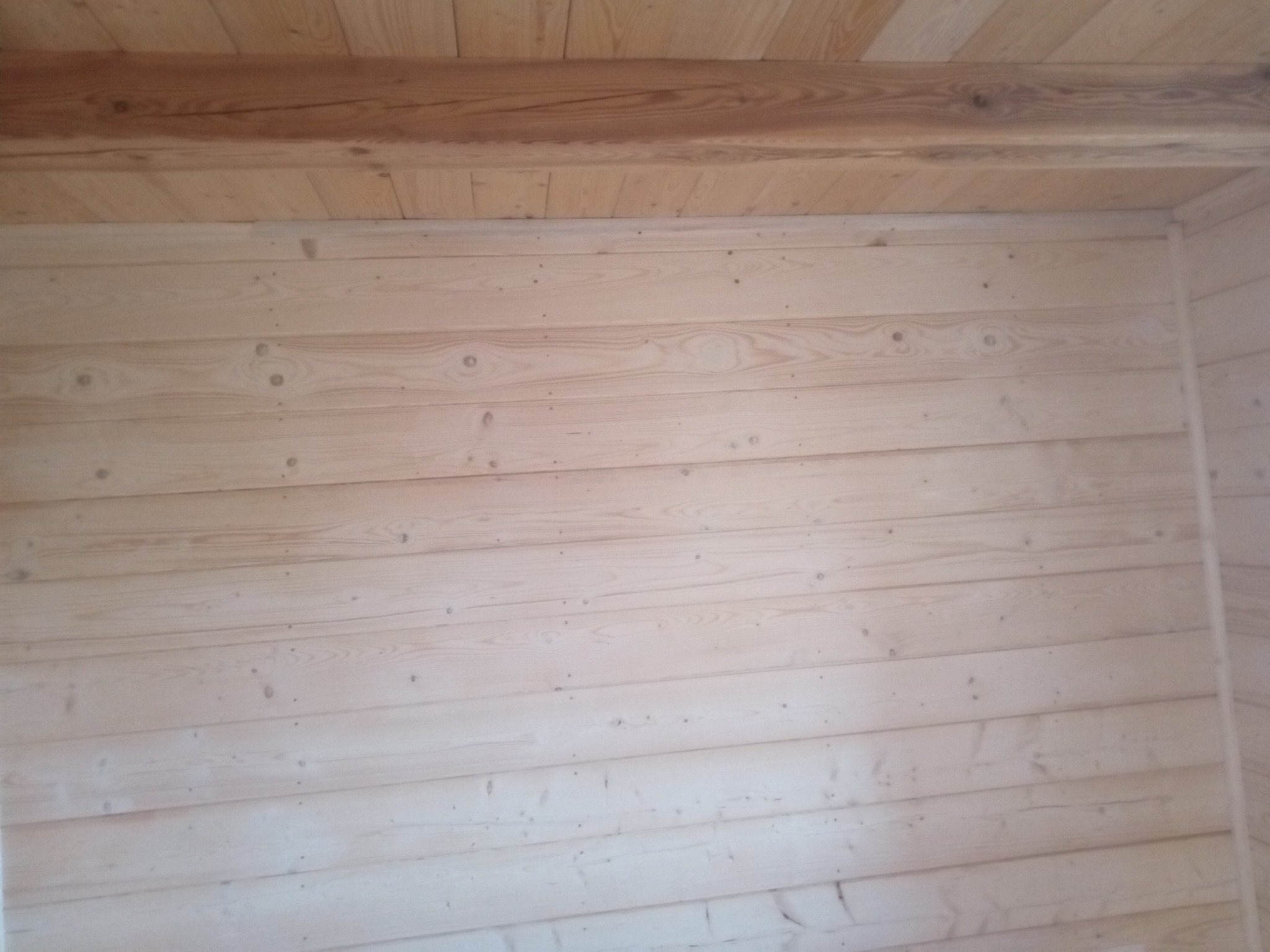 Ściana i sufit wykończone jasnymi, surowymi deskami boazeryjnymi z widocznymi sękami i naturalnym usłojeniem drewna, z drewnianą belką stropową w górnej części kadru.