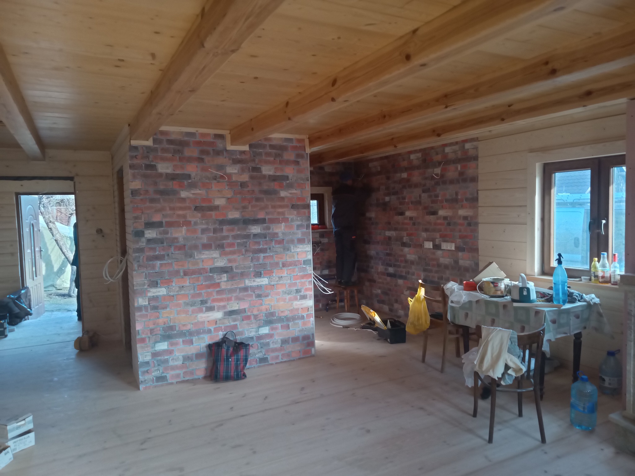 Wnętrze drewnianego domu w trakcie remontu: ściana z cegły dekoracyjnej, drewniana podłoga i sufit, widoczny pracownik na stołku montujący element na ścianie.