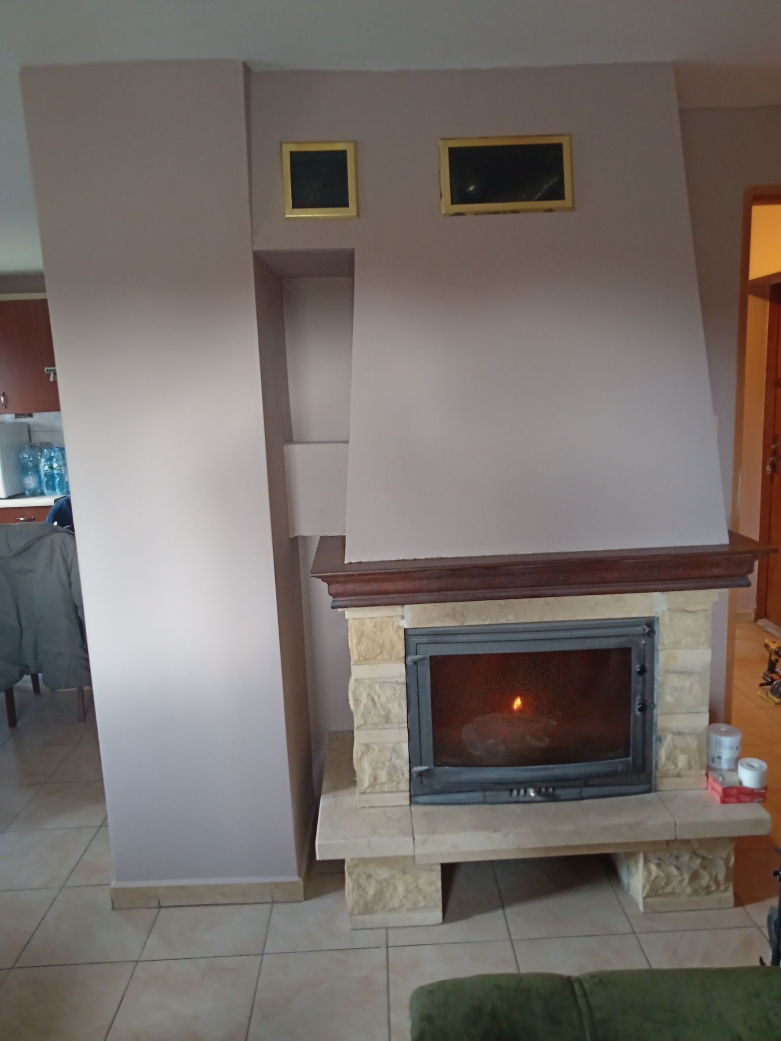 Wnętrze salonu z kominkiem obudowanym kamieniem, drewnianym blatem i elementami dekoracyjnymi w postaci dwóch obrazków w złotych ramach na ścianie pomalowanej na jasny fiolet, widoczne akcesoria...