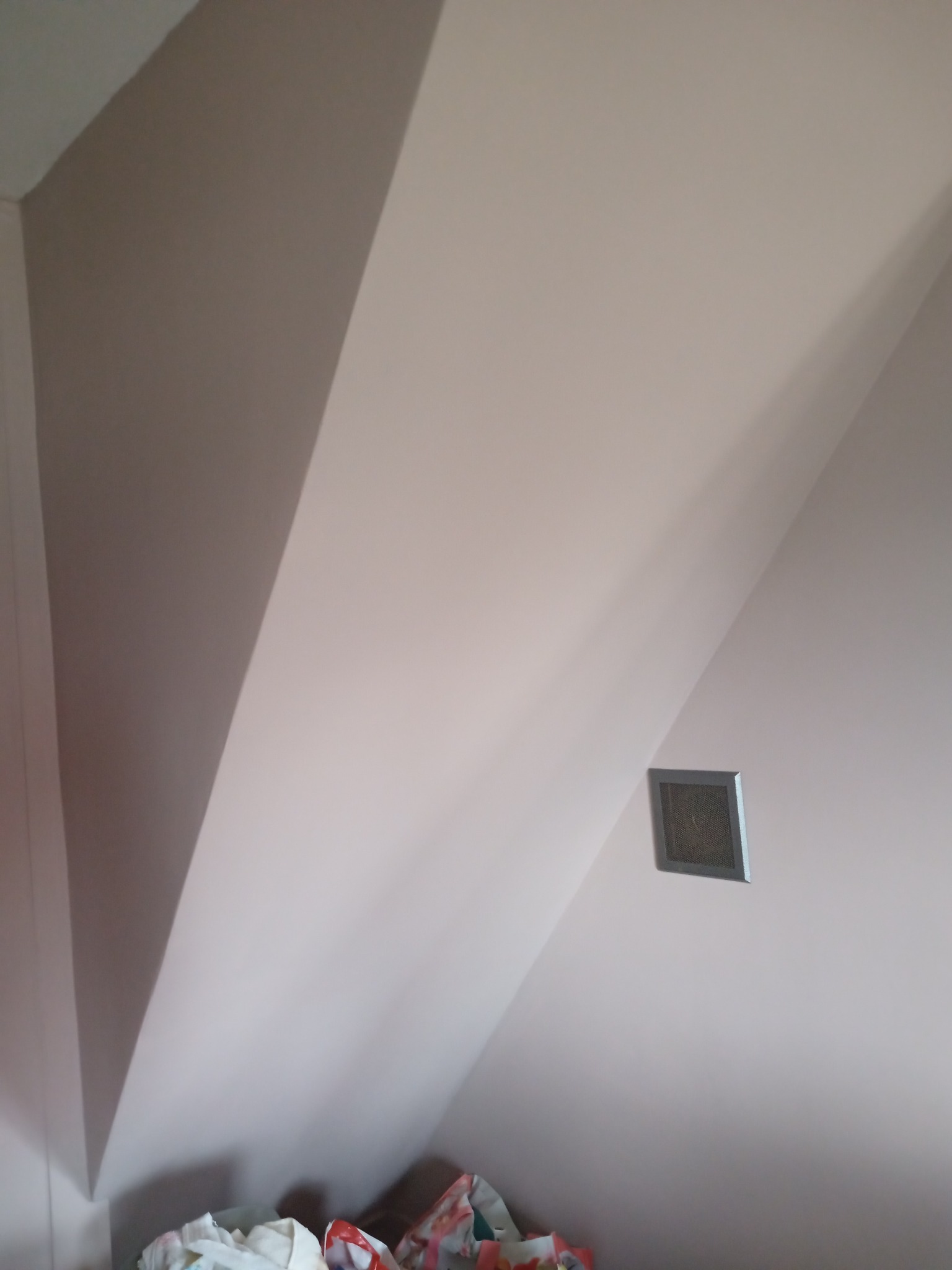 Pomalowane na jasny kolor skosy poddasza z kratką wentylacyjną, widoczne torby z zakupami na dole kadru.