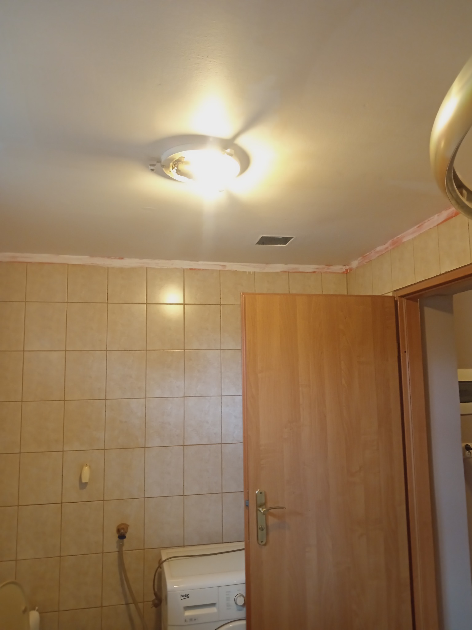 Łazienka w trakcie prac wykończeniowych: widoczne płytki ceramiczne, biała pralka Beko, drewniane drzwi z klamką oraz sufit z lampą i śladami po szpachlowaniu przy łączeniu ze ścianą.