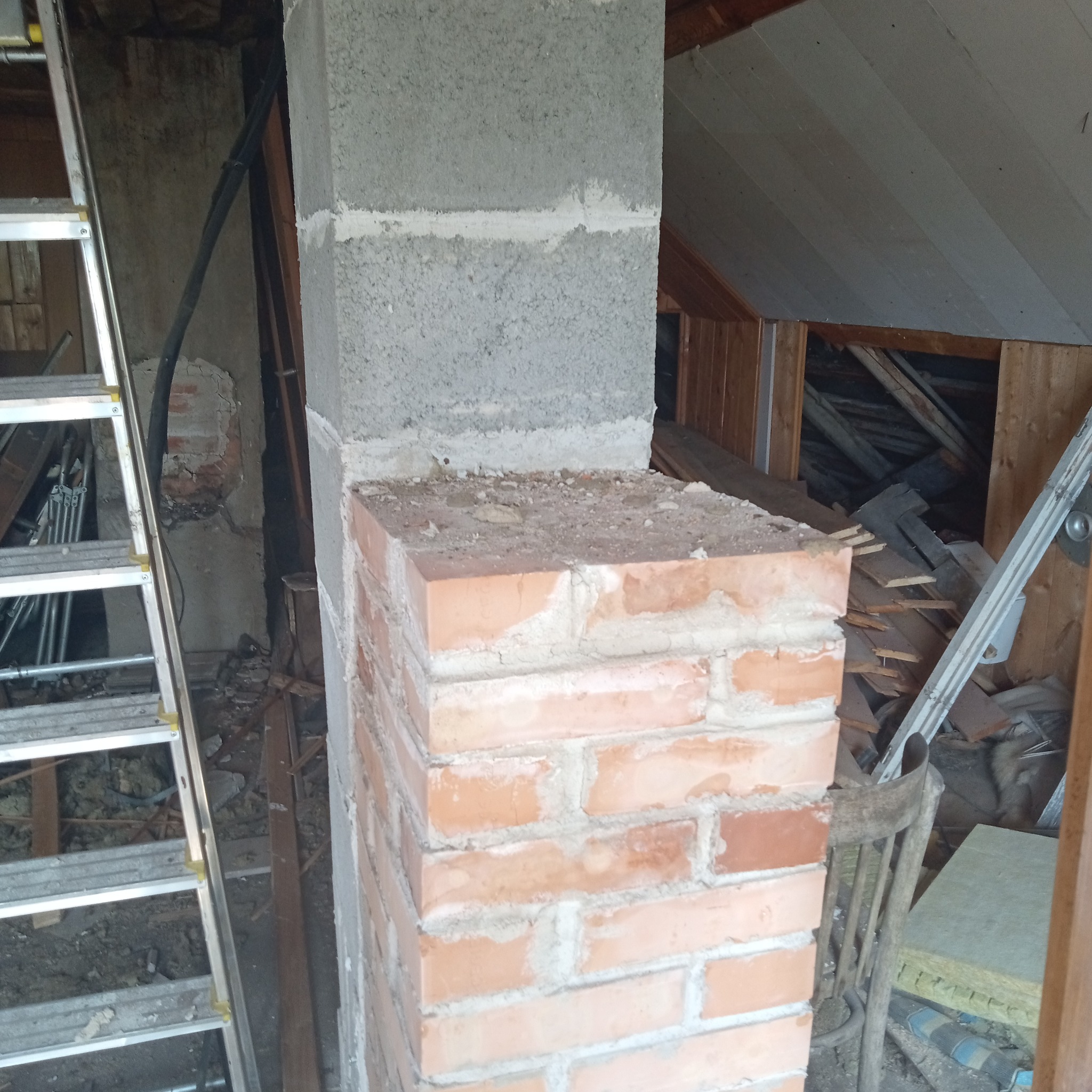 Wnętrze poddasza w trakcie remontu z widocznym kominem z cegły i bloczków betonowych, drabina aluminiowa oparta o ścianę, elementy konstrukcyjne dachu w tle.