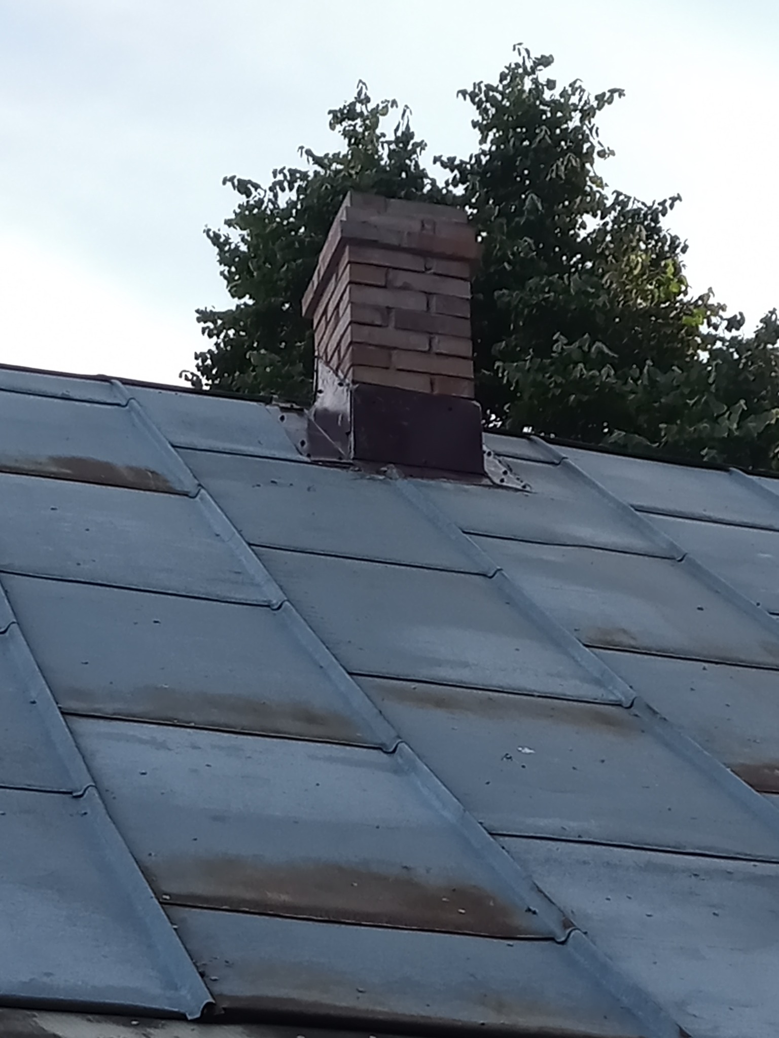 Metalowy dach z ceglanym kominem w tle zielone drzewa i jasne niebo.