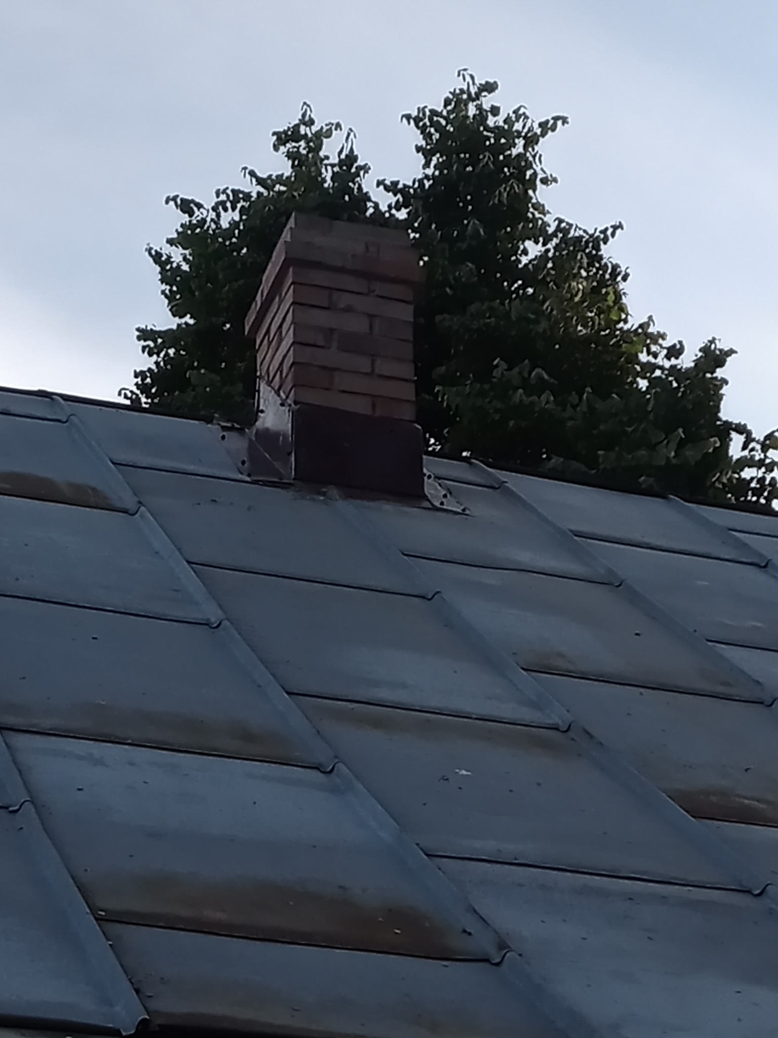 Ceglany komin wystający z metalowego dachu, z zielonymi drzewami w tle i błękitnym niebem.