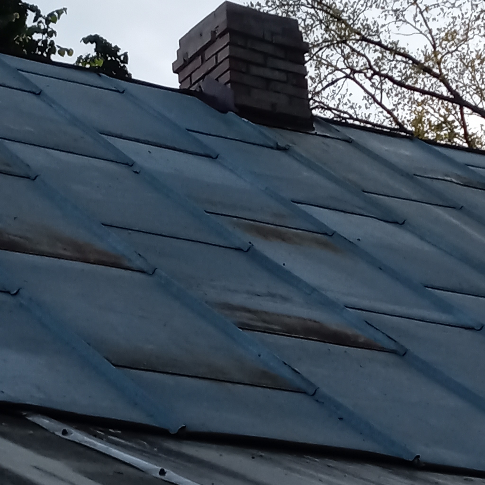 Metalowy dach z ceglanym kominem, widok z dołu, fragment drzewa w tle.