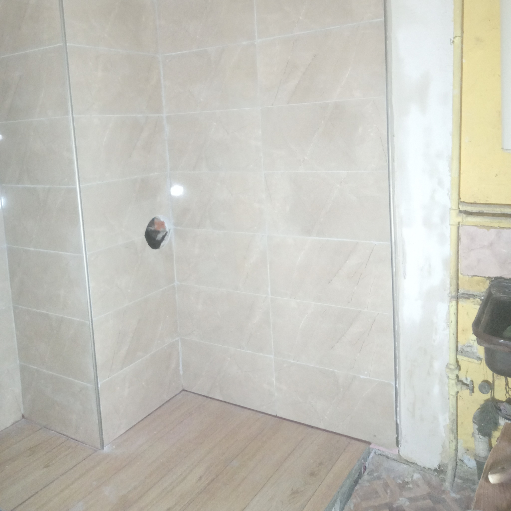 Wykończona narożna kabina prysznicowa z beżowymi płytkami imitującymi marmur, widoczny otwór na armaturę, jasne panele podłogowe oraz fragment starej instalacji wodnej i ściany przed remontem.