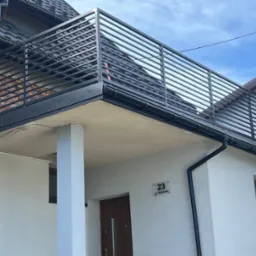 Nowoczesna balustrada balkonowa z poziomych, czarnych profili zamontowana na budynku z białą elewacją, widoczny numer domu i kamera monitoringu.