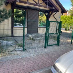 NEX-STAL Łukasz Kurowski - Zielone ogrodzenie panelowe z furtką przed budynkiem z roletami zewnętrznymi, widoczne chwasty i trawa na chodniku, fragment samochodu w prawym dolnym rogu.