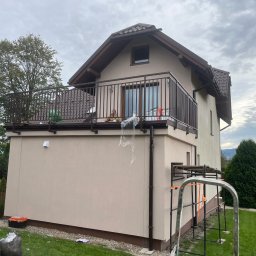 NEX-STAL Łukasz Kurowski - Balkon w nowym domu z brązową balustradą, widok z dołu na tle pochmurnego nieba, widoczne narzędzia i resztki folii ochronnej po montażu.