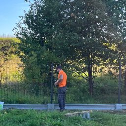 NEX-STAL Łukasz Kurowski - Pracownik w pomarańczowej koszulce montuje ogrodzenie panelowe, używając wkrętarki, widoczne słupki i podmurówka, w tle drzewo i trawiasta skarpa.