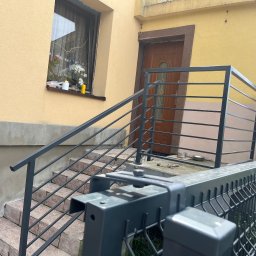 NEX-STAL Łukasz Kurowski - Szare, metalowe balustrady schodowe prowadzące do drzwi wejściowych domu z żółtą elewacją, widok zza przęsła ogrodzenia panelowego.