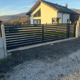 Nowoczesne, poziome ogrodzenie z czarnych paneli na tle domu z jasną elewacją i górskim krajobrazem w oddali.