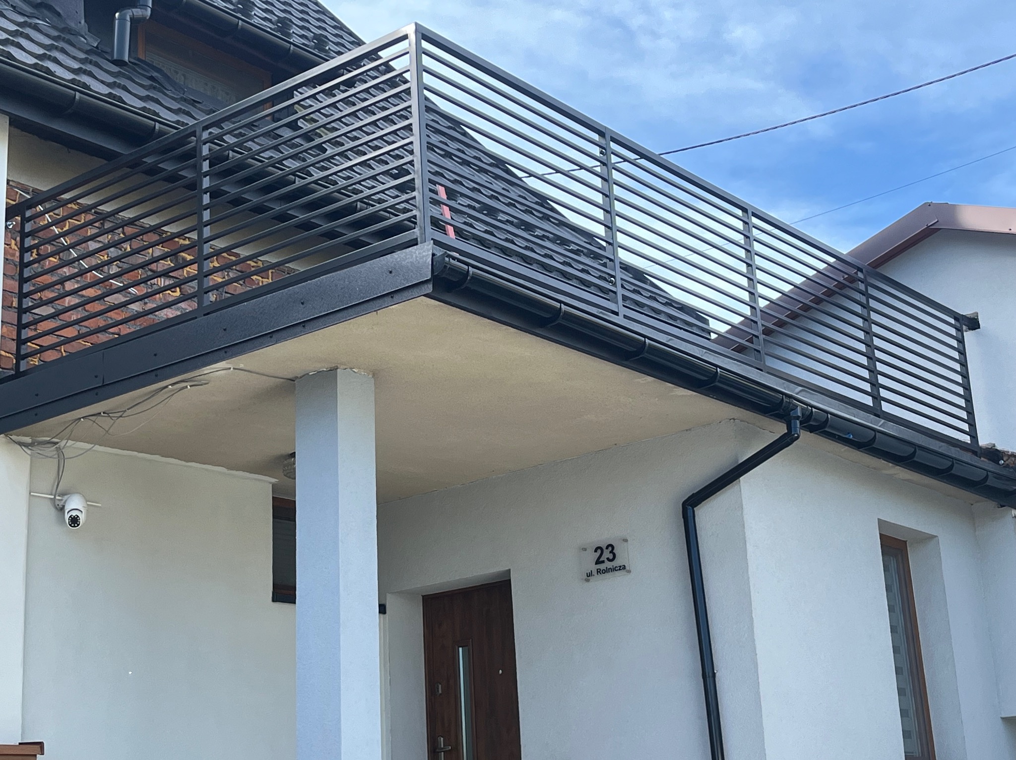 Nowoczesna balustrada balkonowa z poziomych, czarnych profili zamontowana na budynku z białą elewacją, widoczny numer domu i kamera monitoringu.