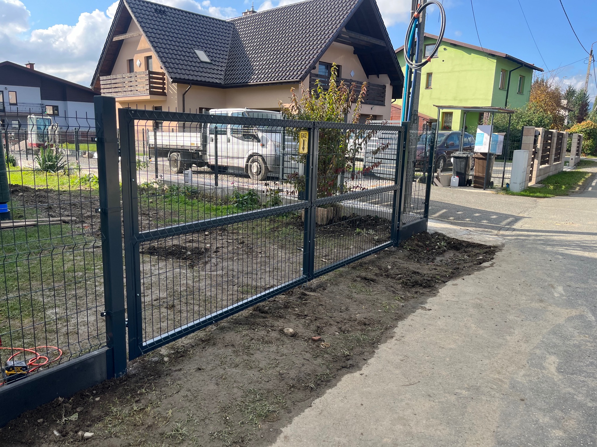 Nowoczesna, ciemnoszara brama panelowa z widocznym znakiem ostrzegawczym 'GAZ' i narzędziami do montażu, na tle domu z beżową elewacją i białym dostawczym samochodem.