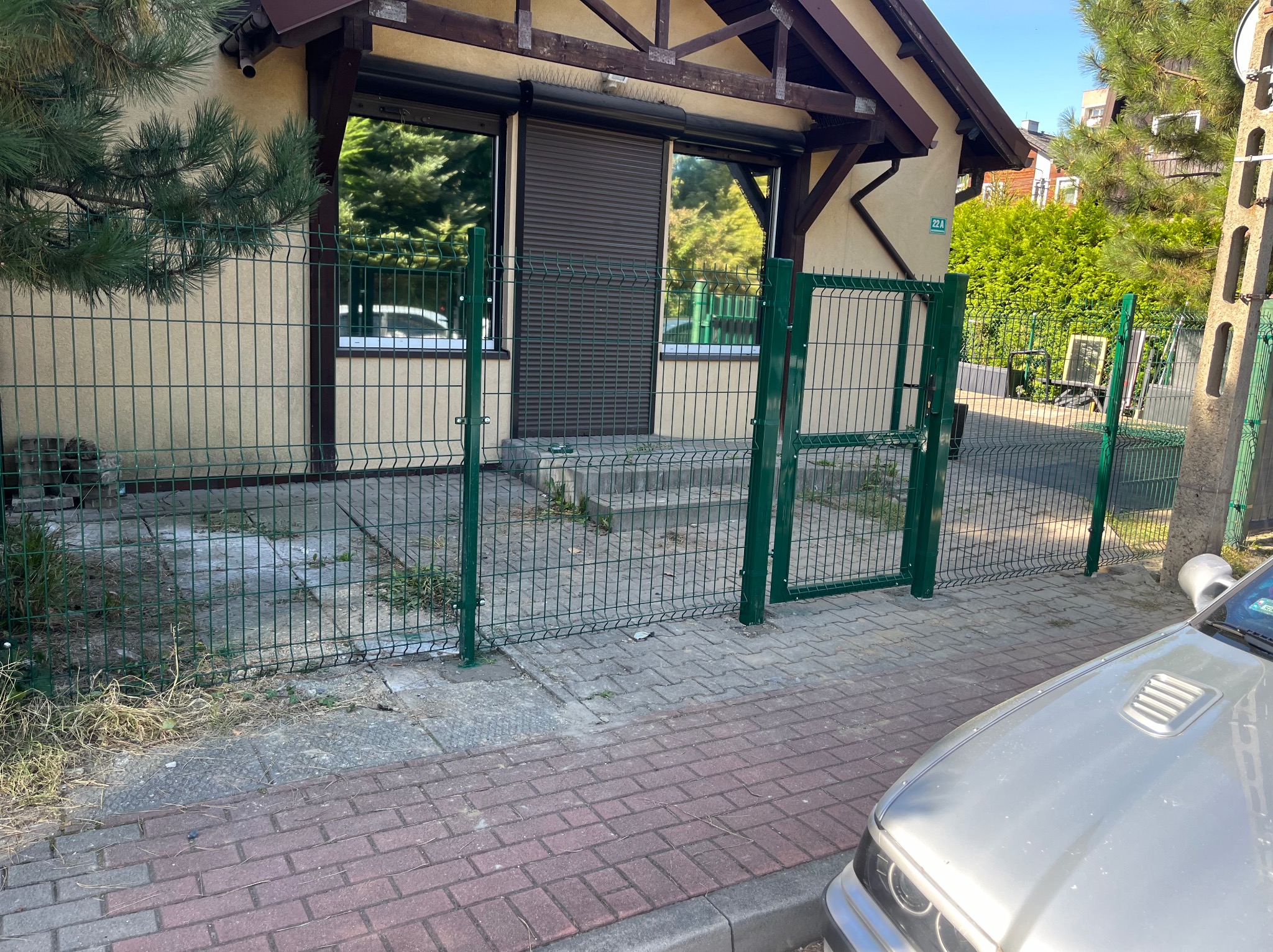 Zielone ogrodzenie panelowe z furtką przed budynkiem z roletami zewnętrznymi, widoczne chwasty i trawa na chodniku, fragment samochodu w prawym dolnym rogu.