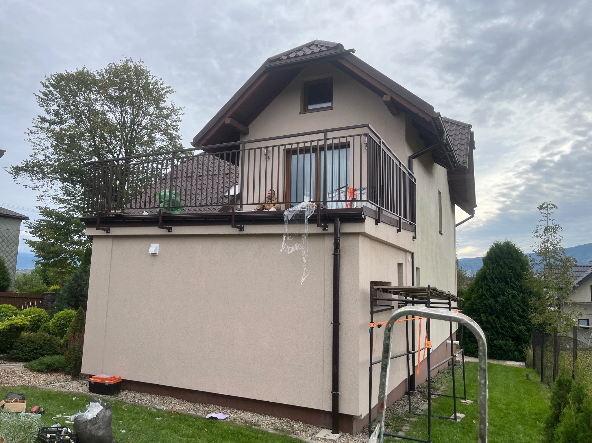 Balkon w nowym domu z brązową balustradą, widok z dołu na tle pochmurnego nieba, widoczne narzędzia i resztki folii ochronnej po montażu.