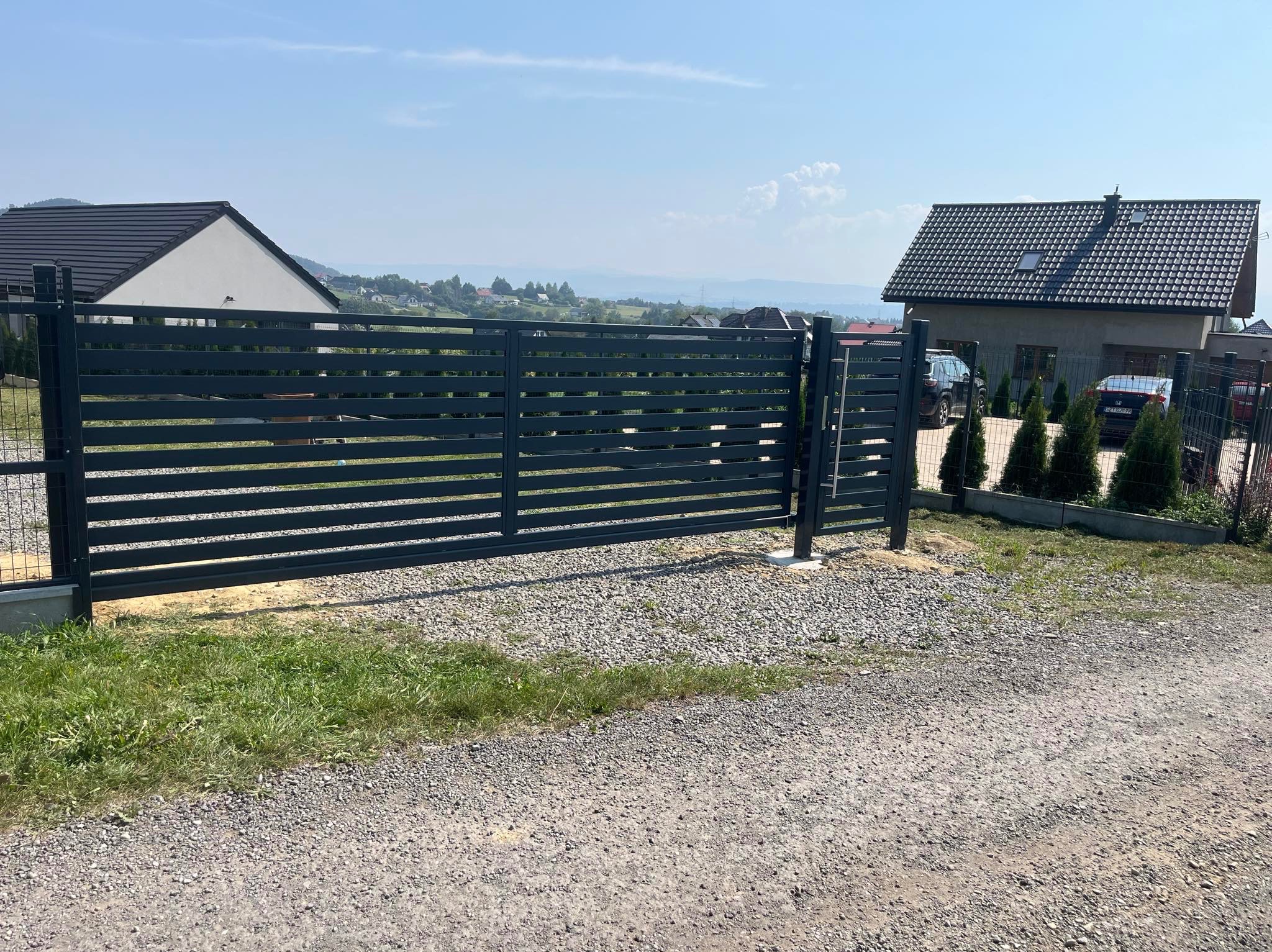 Nowoczesna, pozioma brama wjazdowa z ciemnego metalu, widok z zewnątrz posesji, na tle domów i wzgórz w oddali.