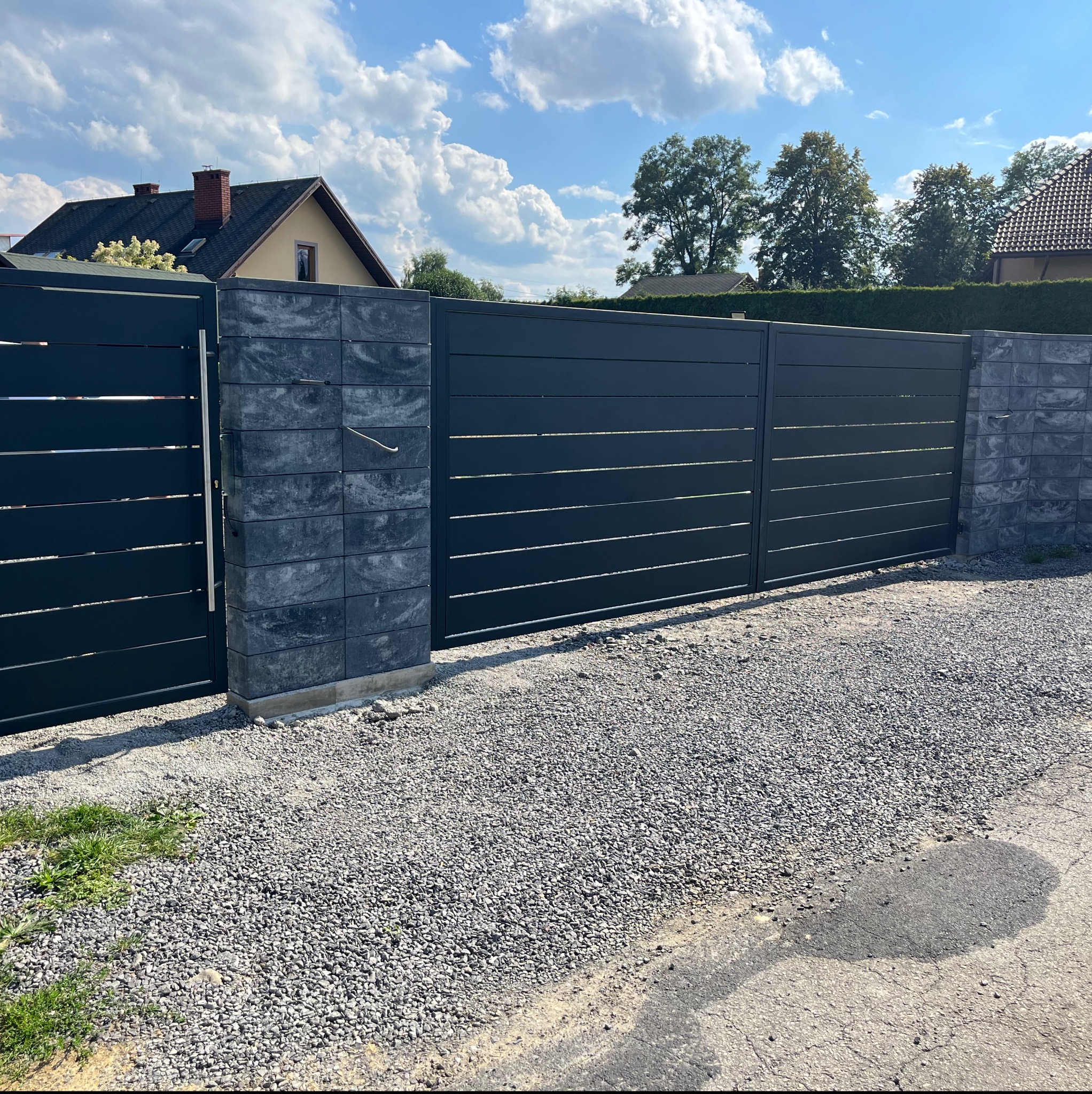 Nowoczesna, pozioma brama wjazdowa w kolorze antracytowym, z metalowymi przęsłami i słupkami z szarej kostki betonowej, na tle błękitnego nieba z białymi chmurami.