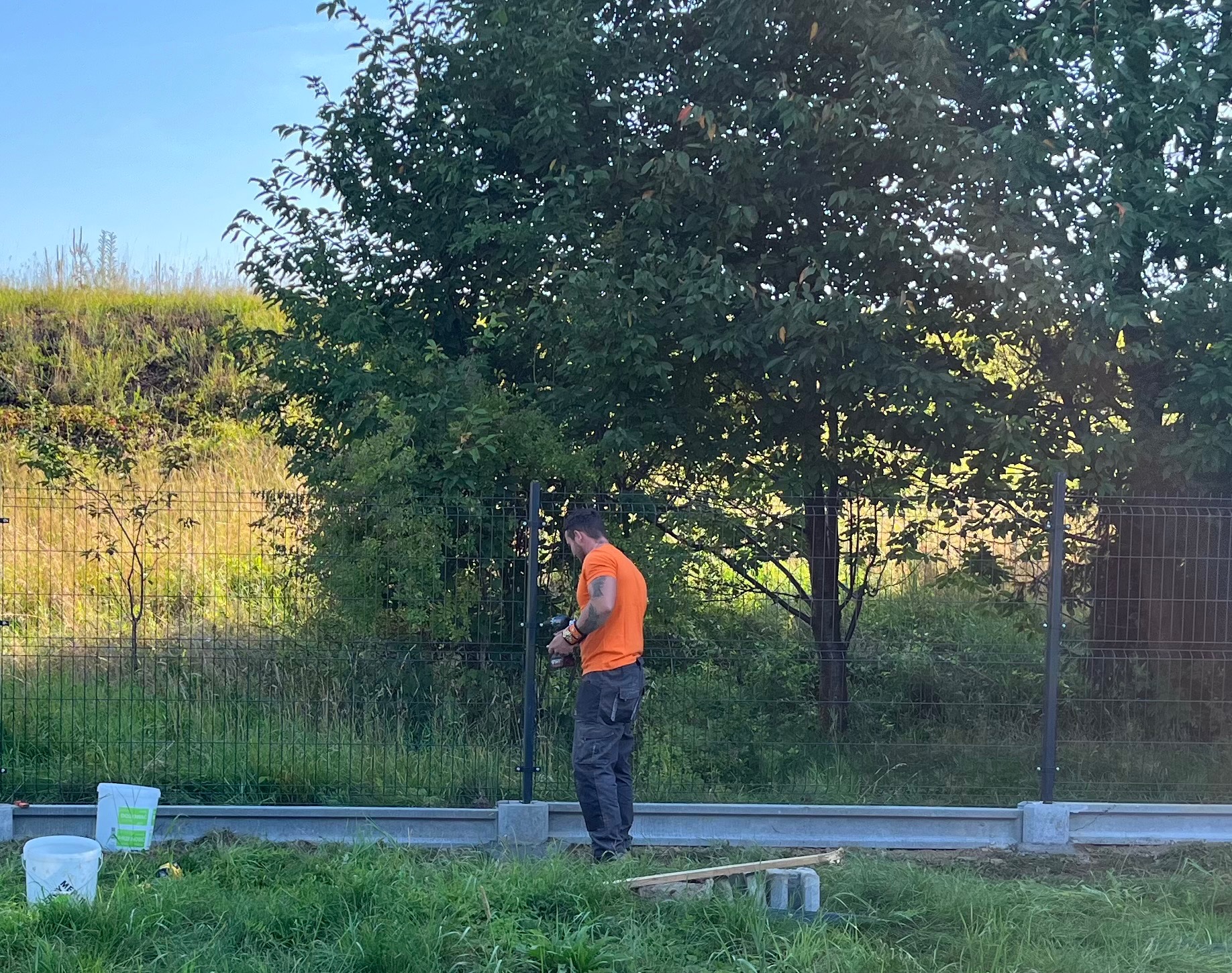 Pracownik w pomarańczowej koszulce montuje ogrodzenie panelowe, używając wkrętarki, widoczne słupki i podmurówka, w tle drzewo i trawiasta skarpa.