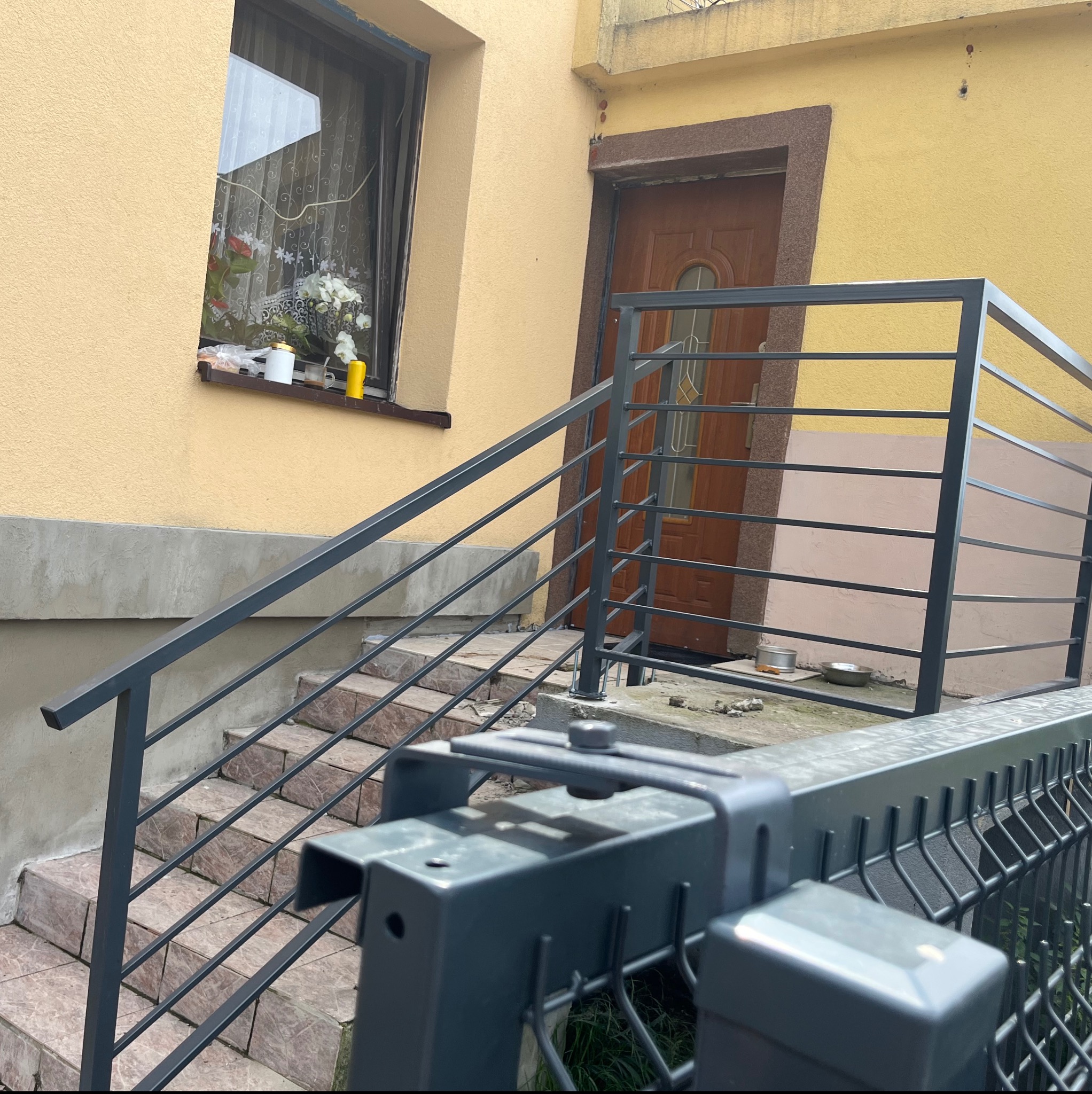 Szare, metalowe balustrady schodowe prowadzące do drzwi wejściowych domu z żółtą elewacją, widok zza przęsła ogrodzenia panelowego.