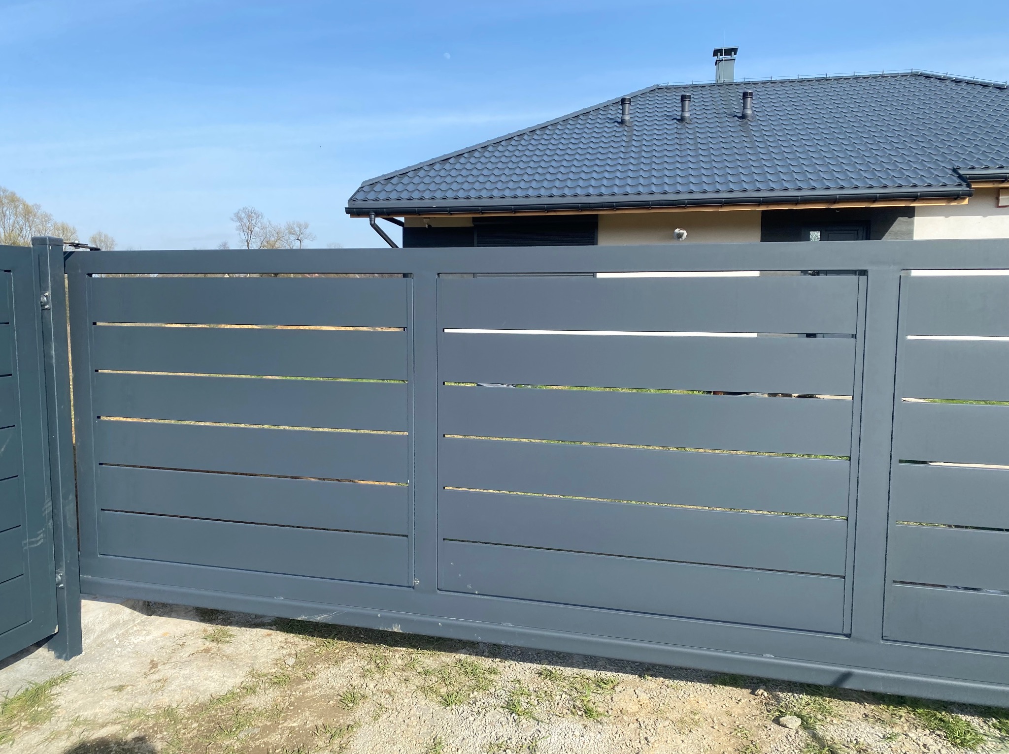 Nowoczesna, przesuwna brama w kolorze grafitowym z poziomymi szczelinami, na tle domu z ciemnoszarą dachówką i błękitnego nieba.