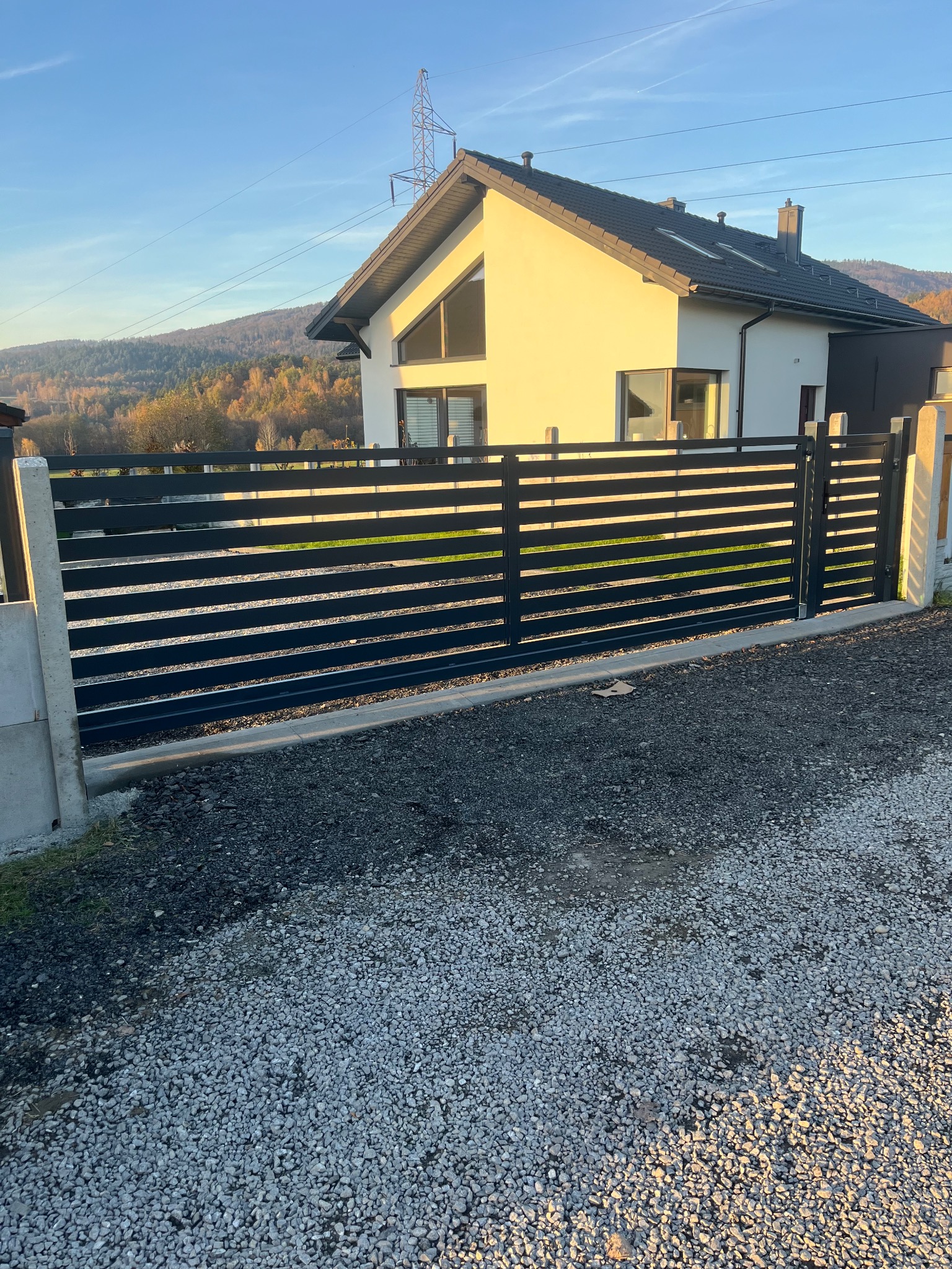 Nowoczesne, poziome ogrodzenie z czarnych paneli na tle domu z jasną elewacją i górskim krajobrazem w oddali.