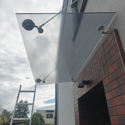 PMOC - Szklany daszek nad wejściem do domu, zamontowany na metalowych wspornikach, widoczna drabina oparta o ścianę budynku.