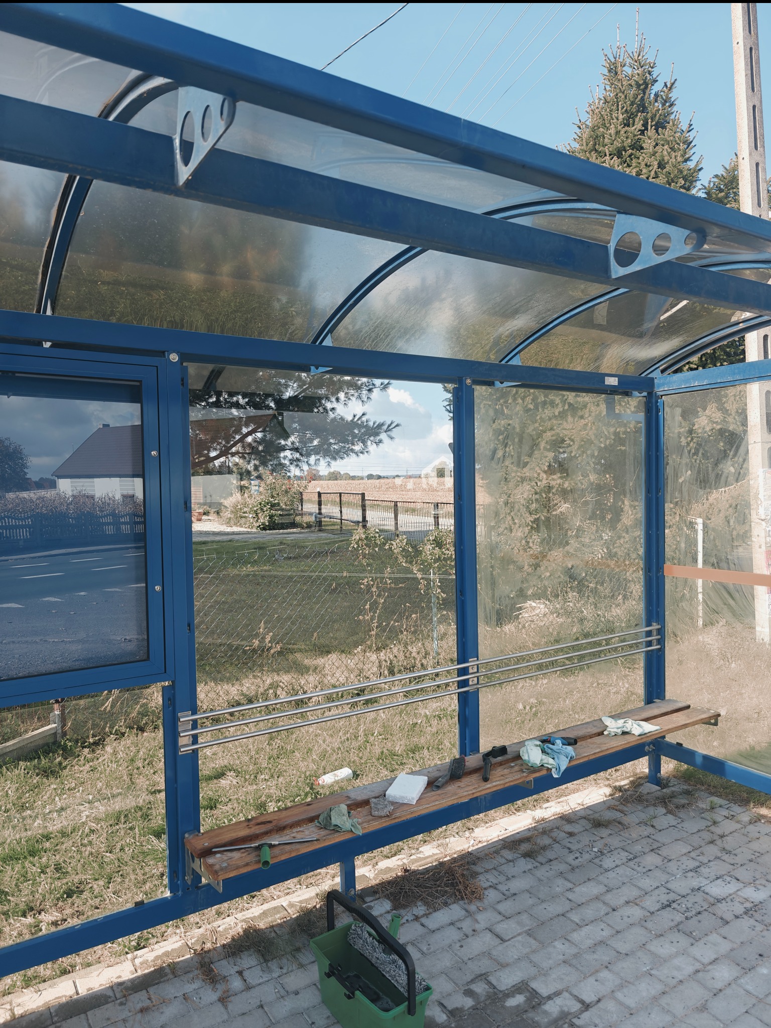 Przystanek autobusowy z nieumytymi szybami, na ławce leżą narzędzia do czyszczenia, widoczny zielony pojemnik z wodą i mopem.