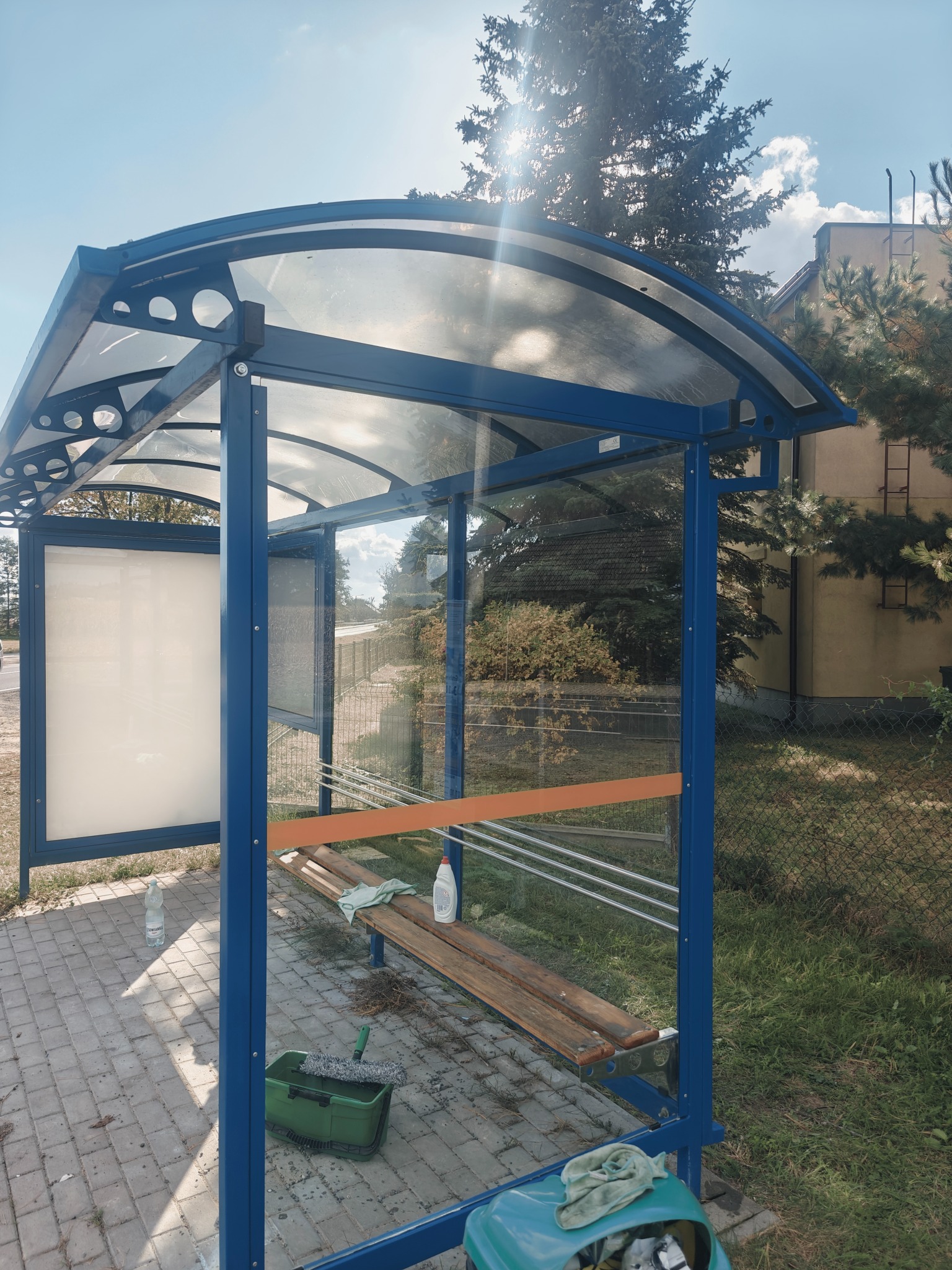 Przystanek autobusowy z niebieską konstrukcją, częściowo umytymi szybami, wiaderkiem, mopem i butelką płynu do mycia szyb na ławce.