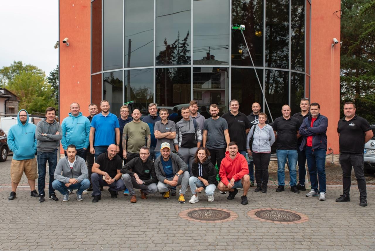 Grupa pracowników firmy sprzątającej okna pozuje przed budynkiem z dużymi oknami, na których widoczny jest sprzęt do mycia okien na długim wysięgniku.