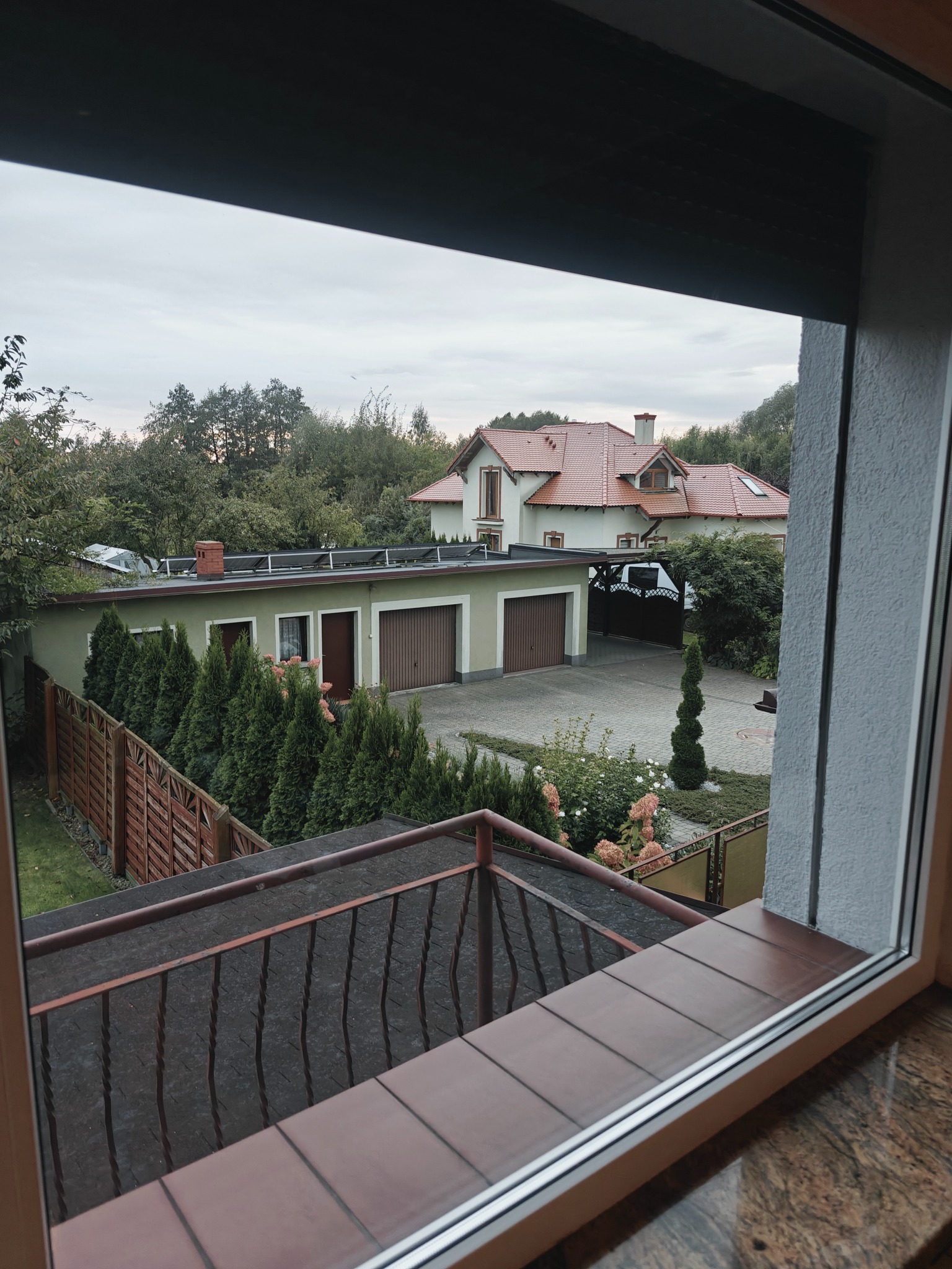 Widok z okna na dom z czerwonym dachem, garaż, zadbany ogród i szpaler iglaków.