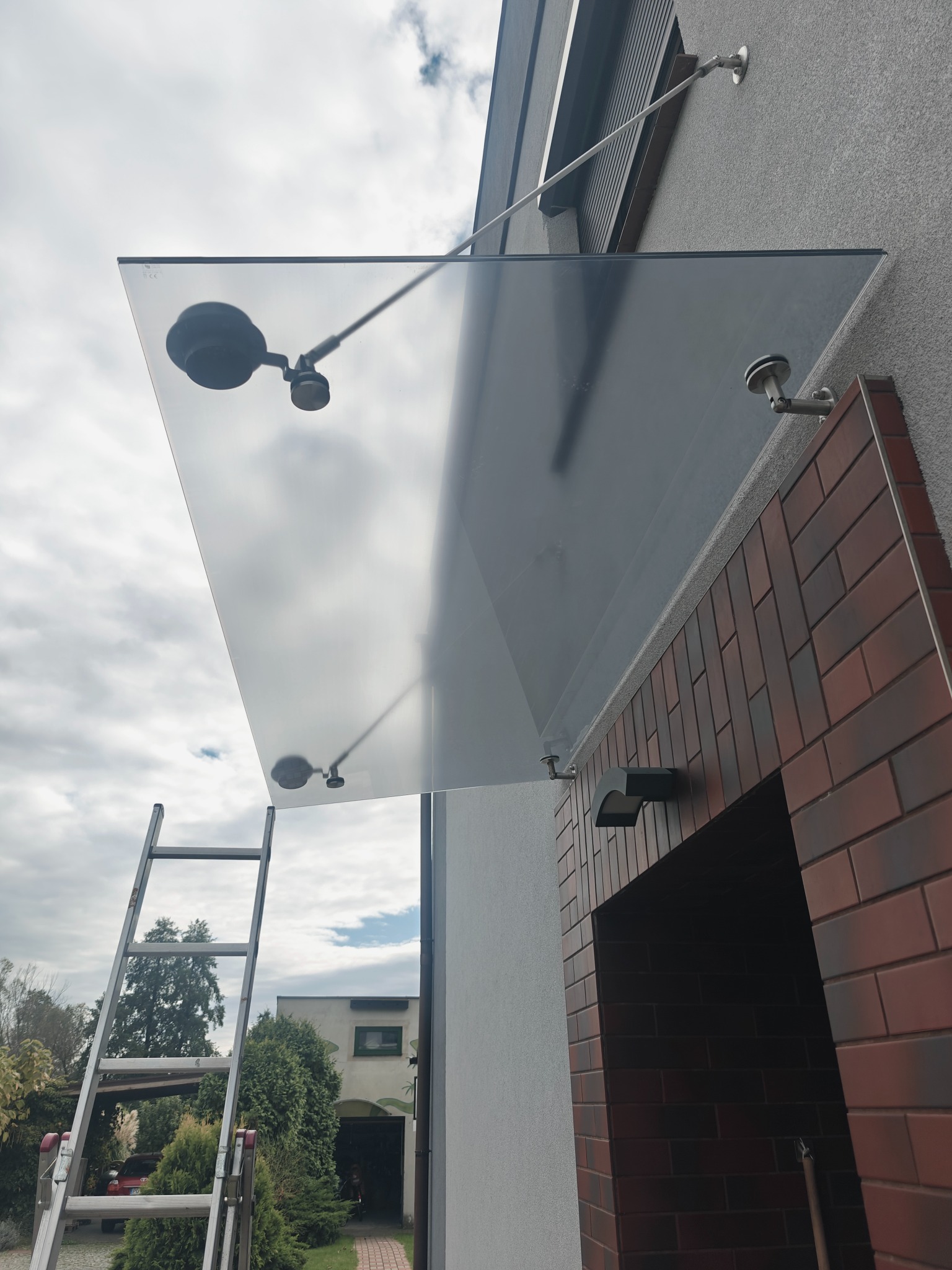 Szklany daszek nad wejściem do domu, zamontowany na metalowych wspornikach, widoczna drabina oparta o ścianę budynku.