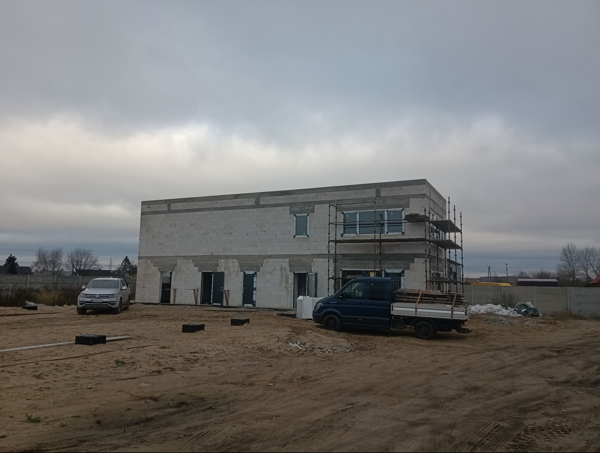 Dwukondygnacyjny budynek w stanie surowym otwartym, wzniesiony z białych bloczków, z widocznymi rusztowaniami, oknami i otworami drzwiowymi, na niezagospodarowanej działce z zaparkowanym samochodem...