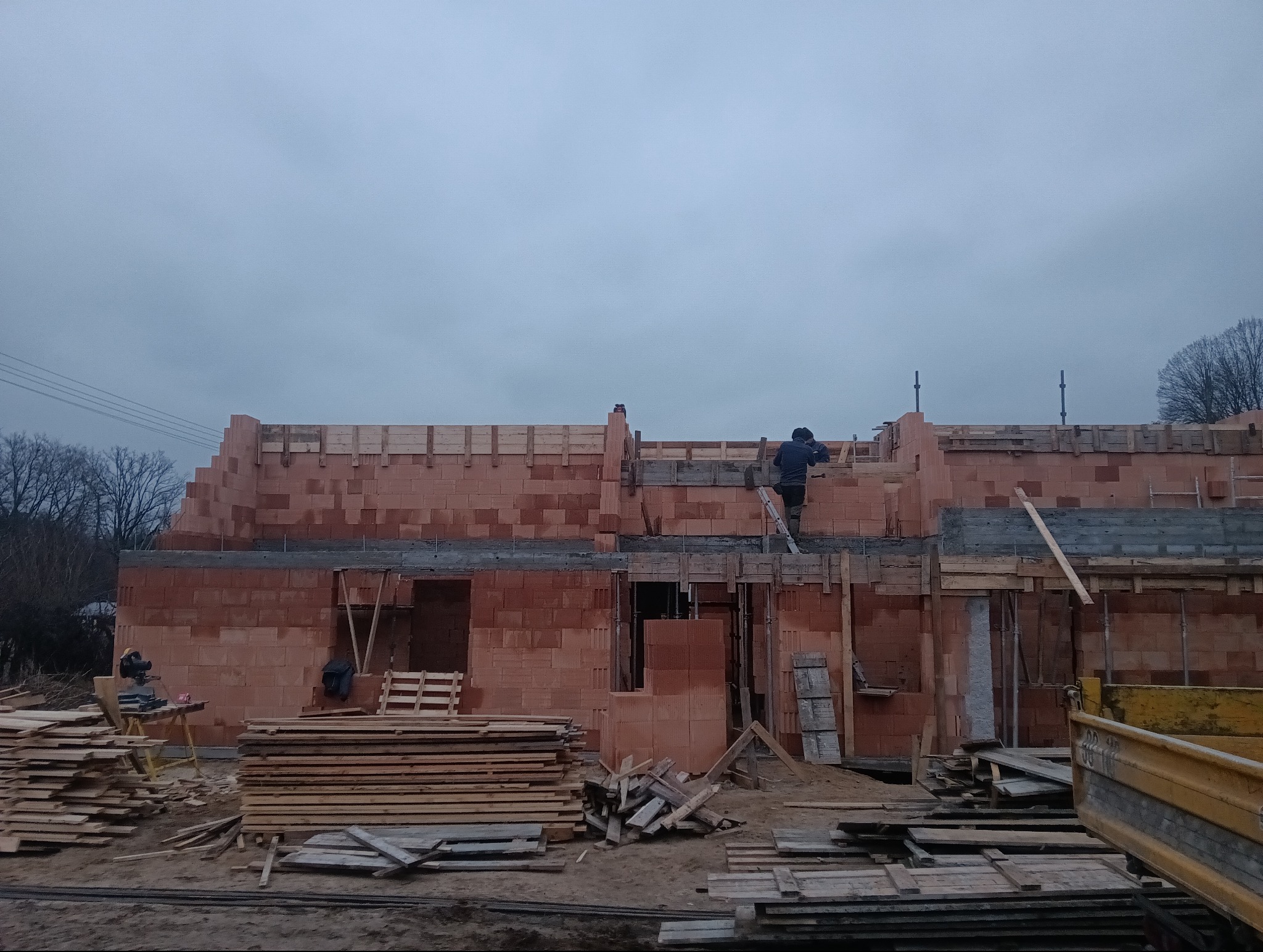 Budowa domu z czerwonej cegły: murarz na drabinie układa ostatnie elementy, widoczne drewniane szalunki, stosy desek i sprzęt budowlany na placu budowy w pochmurny dzień.