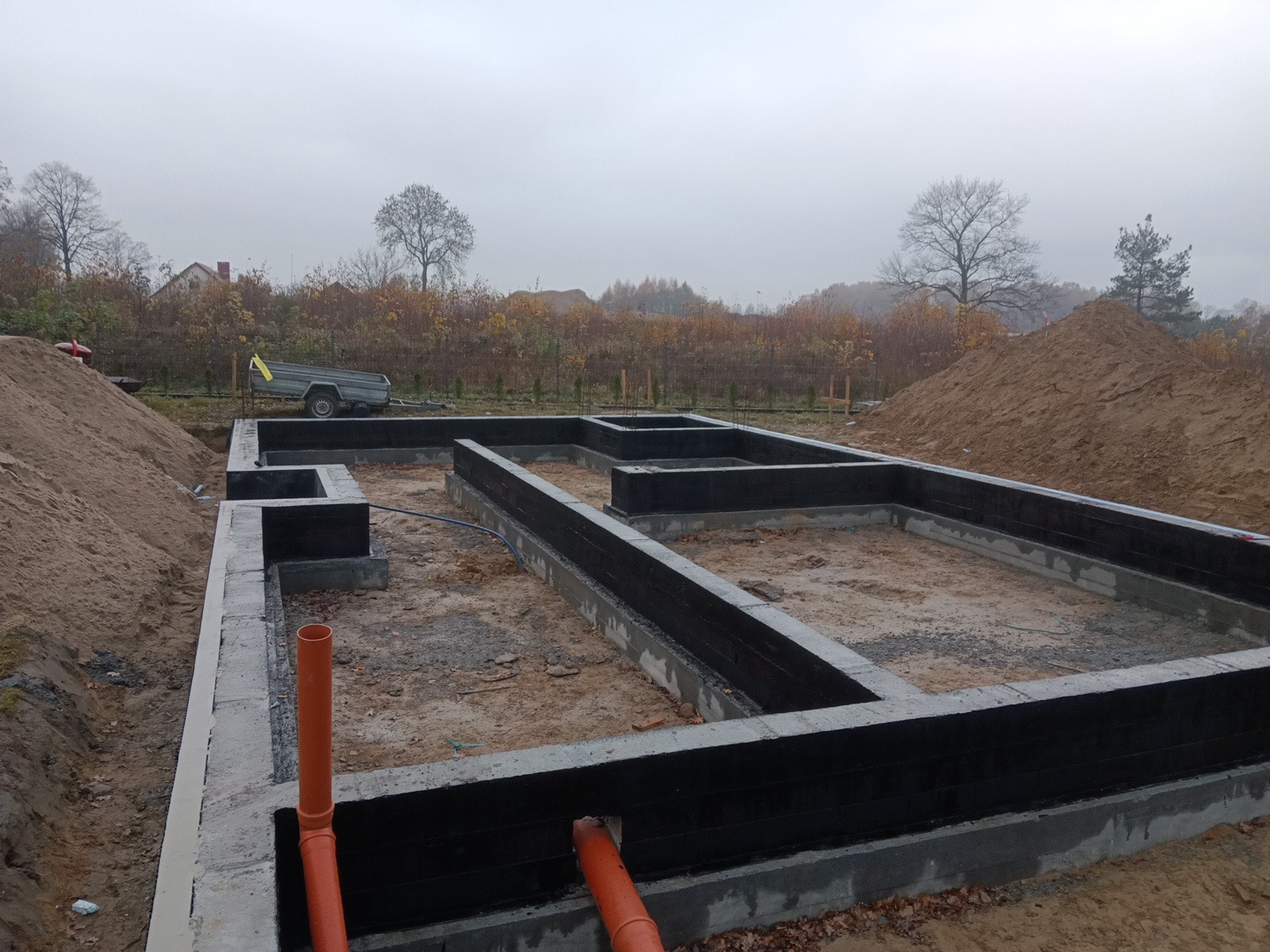 Zalane betonowe fundamenty domu jednorodzinnego z izolacją bitumiczną, widoczne rury kanalizacyjne i wykop pod fundament, w tle przyczepka i hałda ziemi.